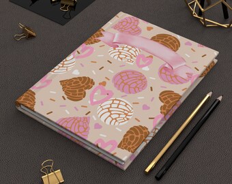 May include: A closed journal with a beige cover featuring a pattern of brown and pink concha bread, hearts, and sprinkles. A pink ribbon is attached. Gold and black pencils and gold binder clips are also present.