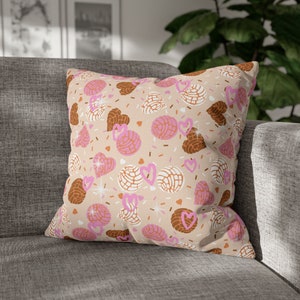 May include: Square decorative pillow with a beige background featuring a repeating pattern of pink and brown conchas, hearts, and sprinkles. The pillow is resting on a gray sofa.