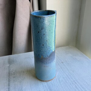 May include: A tall, cylindrical ceramic vase in shades of blue. The vase has a light blue base that fades into a darker, speckled blue towards the top. The inside of the vase is a deep blue.