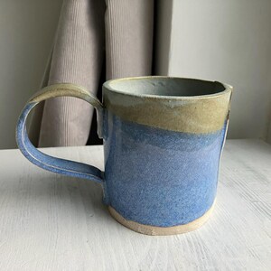 May include: A handcrafted ceramic mug with a large handle. The mug has a blue body with a tan rim and handle. The mug is approximately 10 cm tall and 13 cm wide, suitable for tea or coffee.