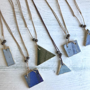 May include: A selection of handmade ceramic pendant necklaces. The pendants are various shapes, including rectangles, squares, and triangles, in shades of blue and green. Each necklace has a natural cord and wooden bead accents.