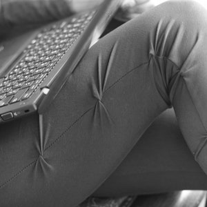 May include: A person is sitting with a laptop computer on their lap. The laptop is open and the keyboard is visible. The person's legs are crossed and their pants are made of a textured fabric.