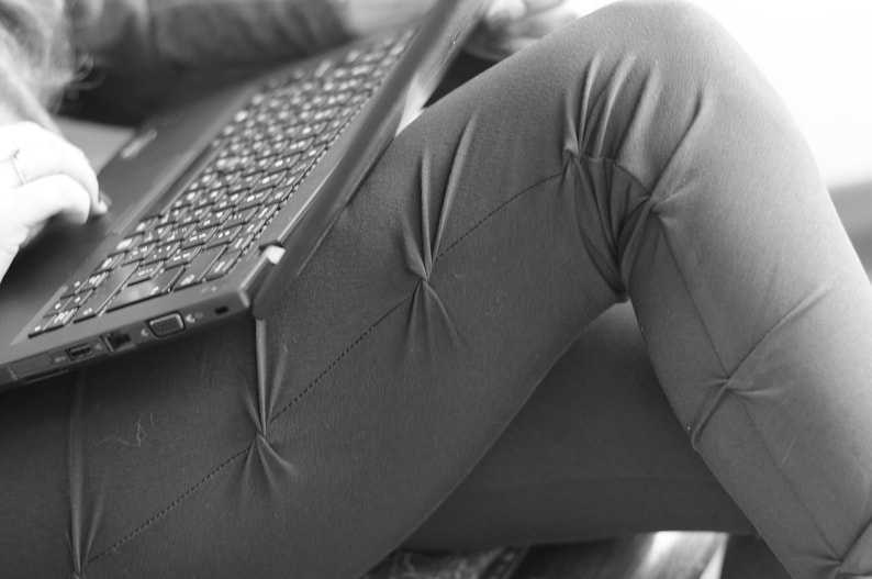 May include: A person is sitting with a laptop computer on their lap. The laptop is open and the keyboard is visible. The person's legs are crossed and their pants are made of a textured fabric.