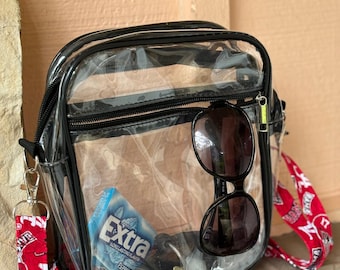 May include: Clear plastic crossbody bag with a black zipper and black trim. The bag has a red and white strap with the text "ALABAMA" on it. The bag contains sunglasses, keys, and a pack of "Extra" peppermint gum.