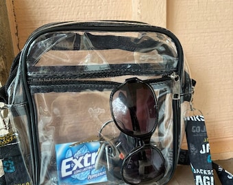 May include: Clear plastic crossbody bag with a black zipper and strap. The bag contains sunglasses, a pack of Extra Peppermint gum, and keys.