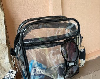 Può includere: Borsa a tracolla trasparente in plastica con cerniera nera e bordatura nera. La borsa ha una tracolla con motivo bianco e nero e contiene occhiali da sole, chewing gum e chiavi.