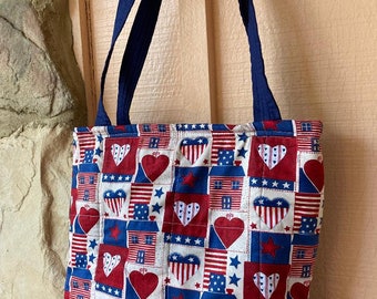 May include: A blue and white quilted tote bag with a red, white and blue patriotic print featuring hearts, stars and houses. The bag has navy blue straps.