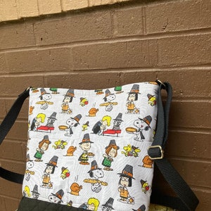 May include: A white and black crossbody bag with a Thanksgiving-themed print featuring cartoon characters. The bag has a black quilted bottom and a black adjustable strap with a gold-colored buckle. The bag is made of fabric.