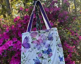 May include: A white tote bag with a purple trim and a floral print featuring butterflies in shades of purple, pink, and blue. The bag is hanging on a metal stand in front of a bush of pink flowers.