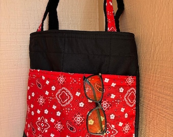 May include: Black tote bag with a red bandana-patterned pocket and black handles. The bag has a quilted design and features a pair of sunglasses in the pocket. The bag is made of fabric.