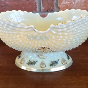 May include: A decorative, pale yellow glass bowl with a scalloped rim and textured, shell-like sides. The bowl sits on a pedestal base with gold and green leaf and shell accents. The rim and base have gold trim.