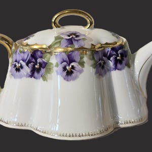 May include: White porcelain teapot with a lid, handle, and spout. The teapot is decorated with purple pansies and gold trim. The handle and lid handle are gold. The base has a scalloped edge.