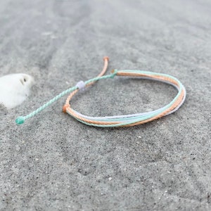 May include: A light blue and peach colored friendship bracelet on a sandy beach.