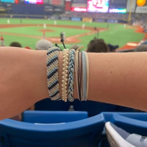 May include: A person's wrist wearing five bracelets. The bracelets are made of different materials, including string, beads, and woven fabric. The bracelets are in shades of blue, white, and black.