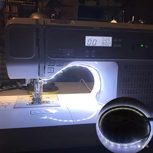 May include: White LED light strip with a clamp for attaching to a sewing machine. The light strip is curved around the arm of the sewing machine, providing bright illumination for sewing projects.