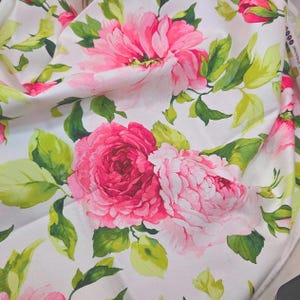 May include: A white fabric featuring a floral pattern with large, layered pink and magenta peony blossoms. The design includes vibrant green leaves and stems scattered throughout the textile, creating a dense, colorful botanical print on a plain background.