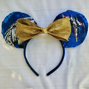 May include: Star Wars themed Minnie Mouse ears headband with a gold glitter bow. The ears are blue with white stars and feature images of the Millennium Falcon and X-Wing.