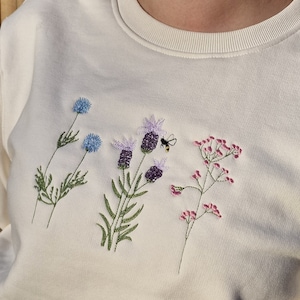 Op de afbeelding: Een wit sweatshirt met geborduurde bloemen en een bij. De bloemen zijn paars, blauw en roze.