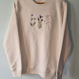 Op de afbeelding: Een cr&egrave;mekleurige trui met geborduurde bloemen in tinten blauw, paars, roze en groen. De bloemen zijn verticaal op de voorkant van de trui gerangschikt.