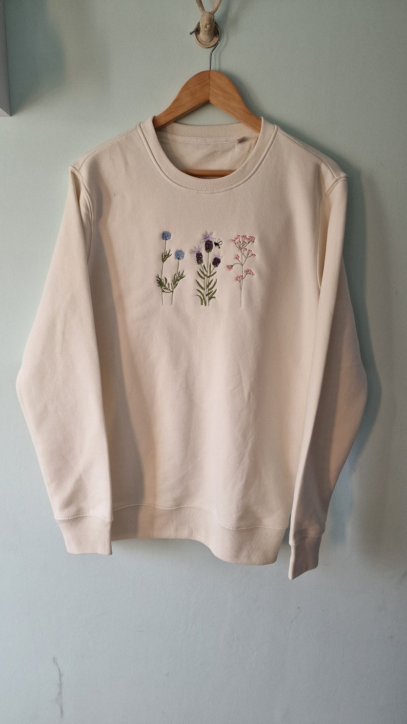 Op de afbeelding: Een cr&egrave;mekleurige trui met geborduurde bloemen in tinten blauw, paars, roze en groen. De bloemen zijn verticaal op de voorkant van de trui gerangschikt.