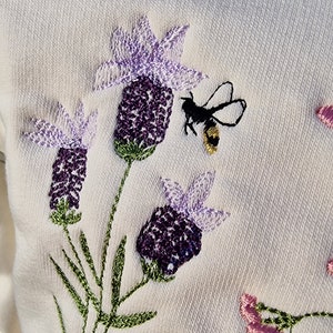 Op de afbeelding: Witte trui met geborduurde lavendelbloemen en een bij. De bloemen zijn paars en lichtpaars met groene stelen en bladeren.