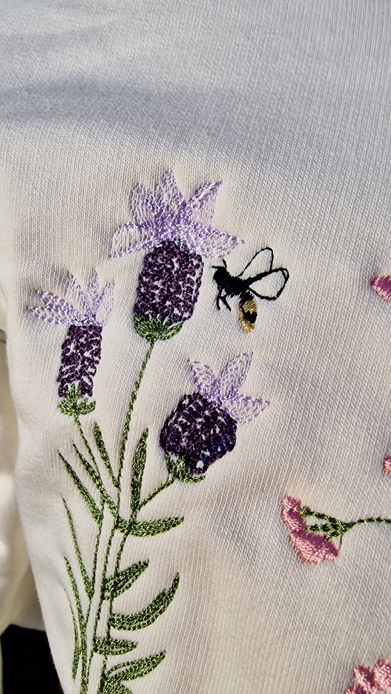 Op de afbeelding: Witte trui met geborduurde lavendelbloemen en een bij. De bloemen zijn paars en lichtpaars met groene stelen en bladeren.