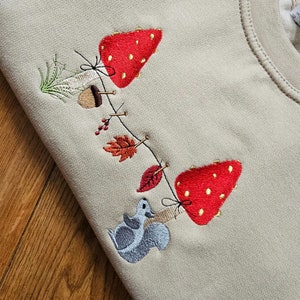 May include: A beige sweatshirt featuring an embroidered design of a squirrel and a string of autumn leaves, mushrooms, and acorns.