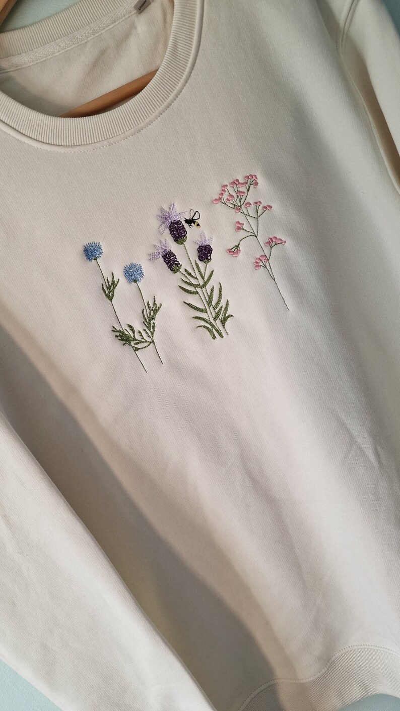 Op de afbeelding: Een wit sweatshirt met geborduurde bloemen en een bij. De bloemen zijn blauw, roze en paars.