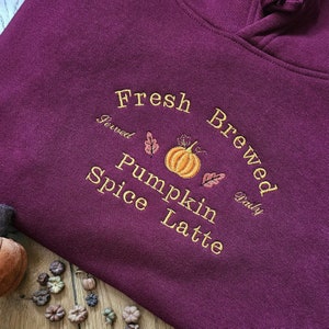May include: A maroon hoodie with the embroidered text "Fresh Brewed Pumpkin Spice Latte." The design includes a pumpkin and leaves in shades of orange and pink. The hoodie is made of a soft material.