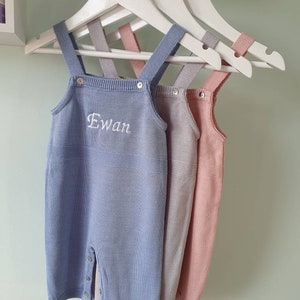May include: Three knitted baby onesies hanging on white hangers. The onesies are blue, gray, and pink. The blue onesie has the name "Ewan" embroidered on the front.