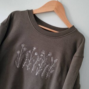 Op de afbeelding: Een donkergroen sweatshirt met een wit geborduurd ontwerp van wilde bloemen.