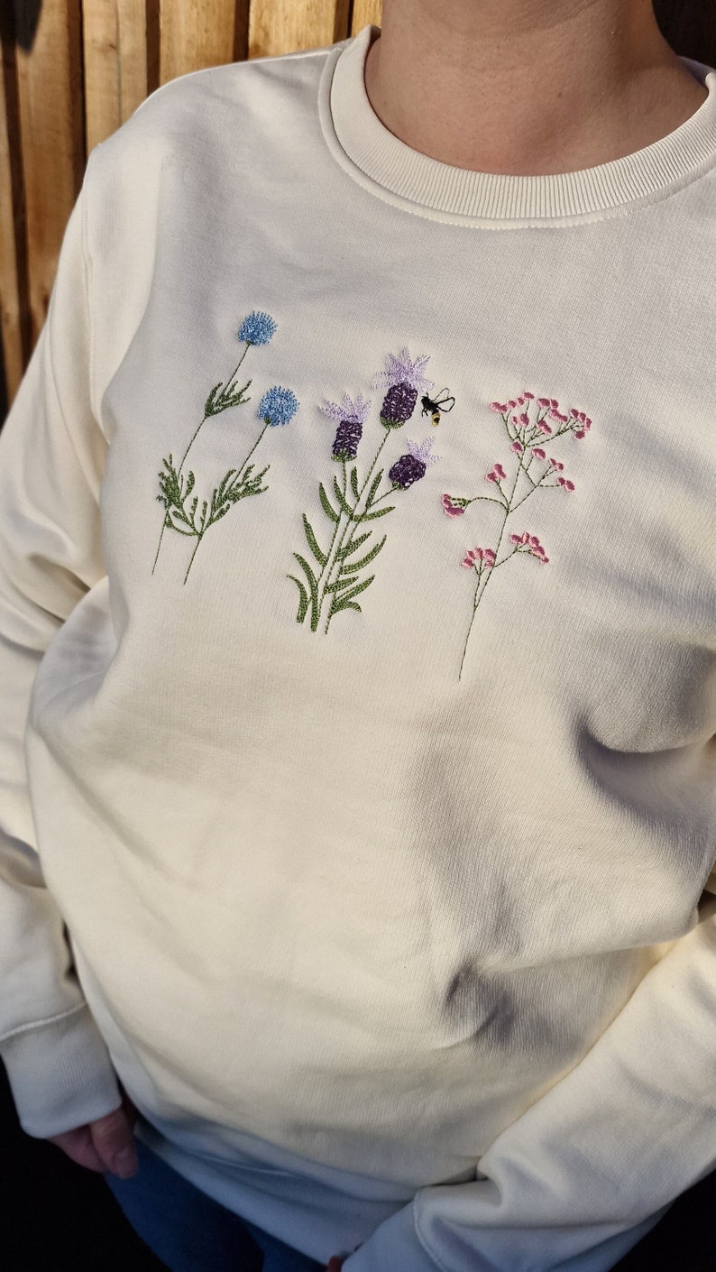 Op de afbeelding: Een wit sweatshirt met geborduurde bloemen en een bij. De bloemen zijn paars, blauw en roze.