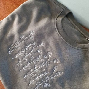 Op de afbeelding: Een grijs sweatshirt met een wit geborduurd ontwerp van een groep bloemen en bladeren.