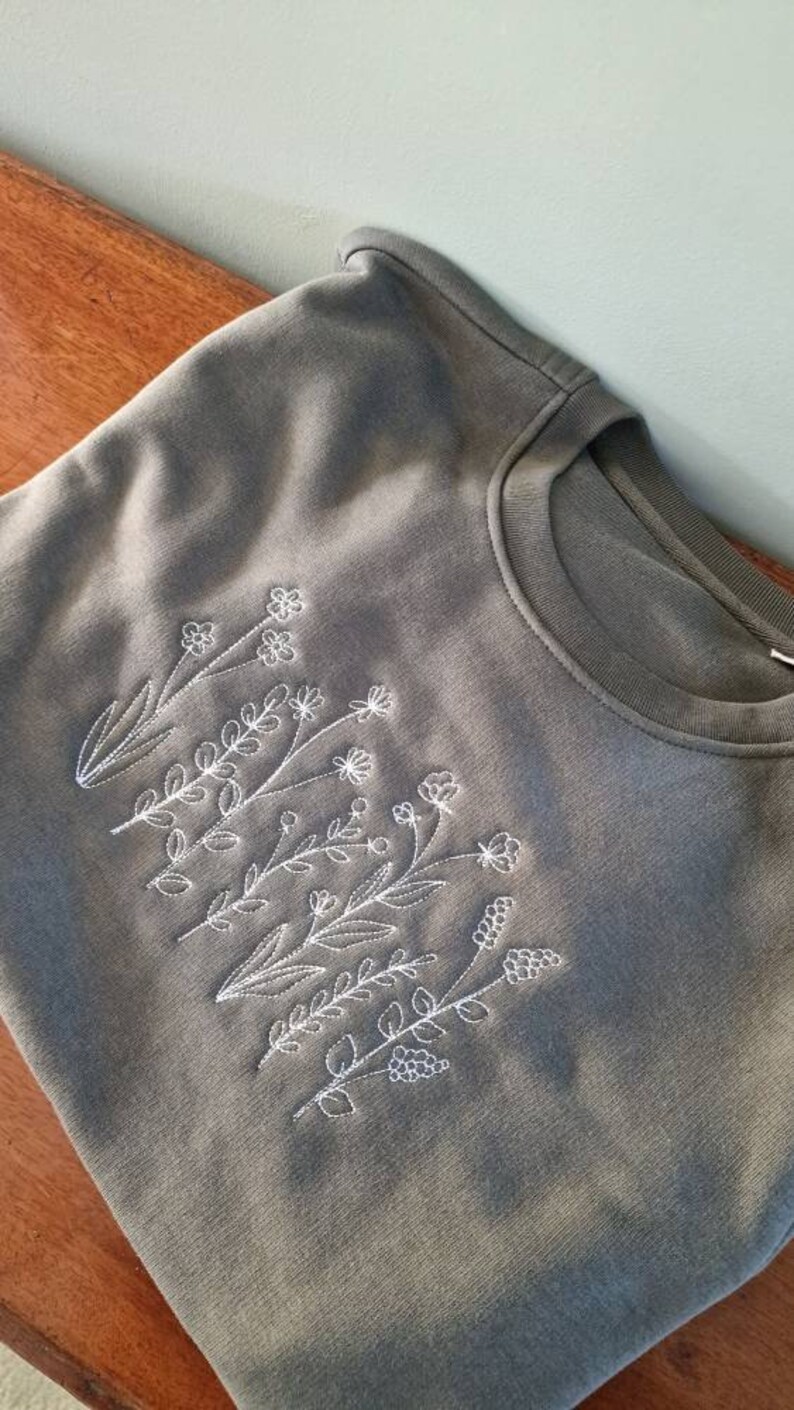 Op de afbeelding: Een grijs sweatshirt met een wit geborduurd ontwerp van een groep bloemen en bladeren.