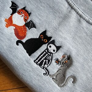 May include: A gray t-shirt with four embroidered cats. One cat is orange and has bat wings. Another cat is black with large eyes. The third cat is a skeleton with a spider. The last cat is gray and has a spider.
