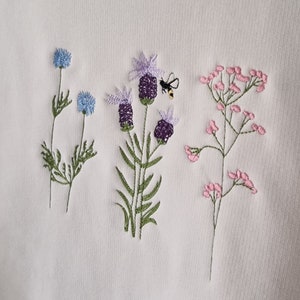 Op de afbeelding: Witte trui met geborduurde bloemen en een bij. De bloemen zijn blauw, paars en roze.