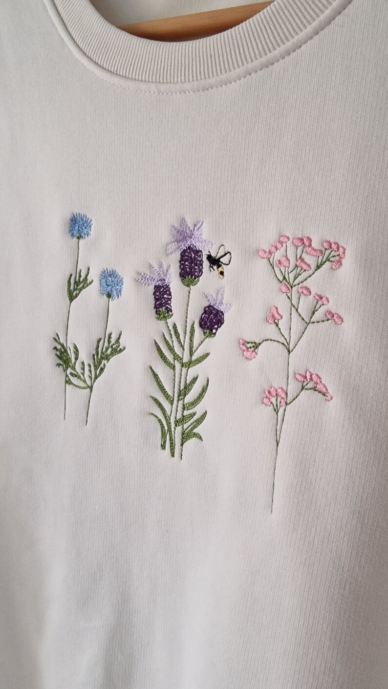 Op de afbeelding: Witte trui met geborduurde bloemen en een bij. De bloemen zijn blauw, paars en roze.