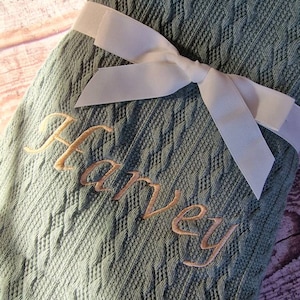 May include: A green cable knit baby blanket with a white satin ribbon tied in a bow. The blanket is personalized with the name "Harvey" embroidered in gold thread.