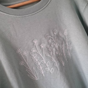 Op de afbeelding: Een lichtblauw sweatshirt met witte geborduurde bloemen.