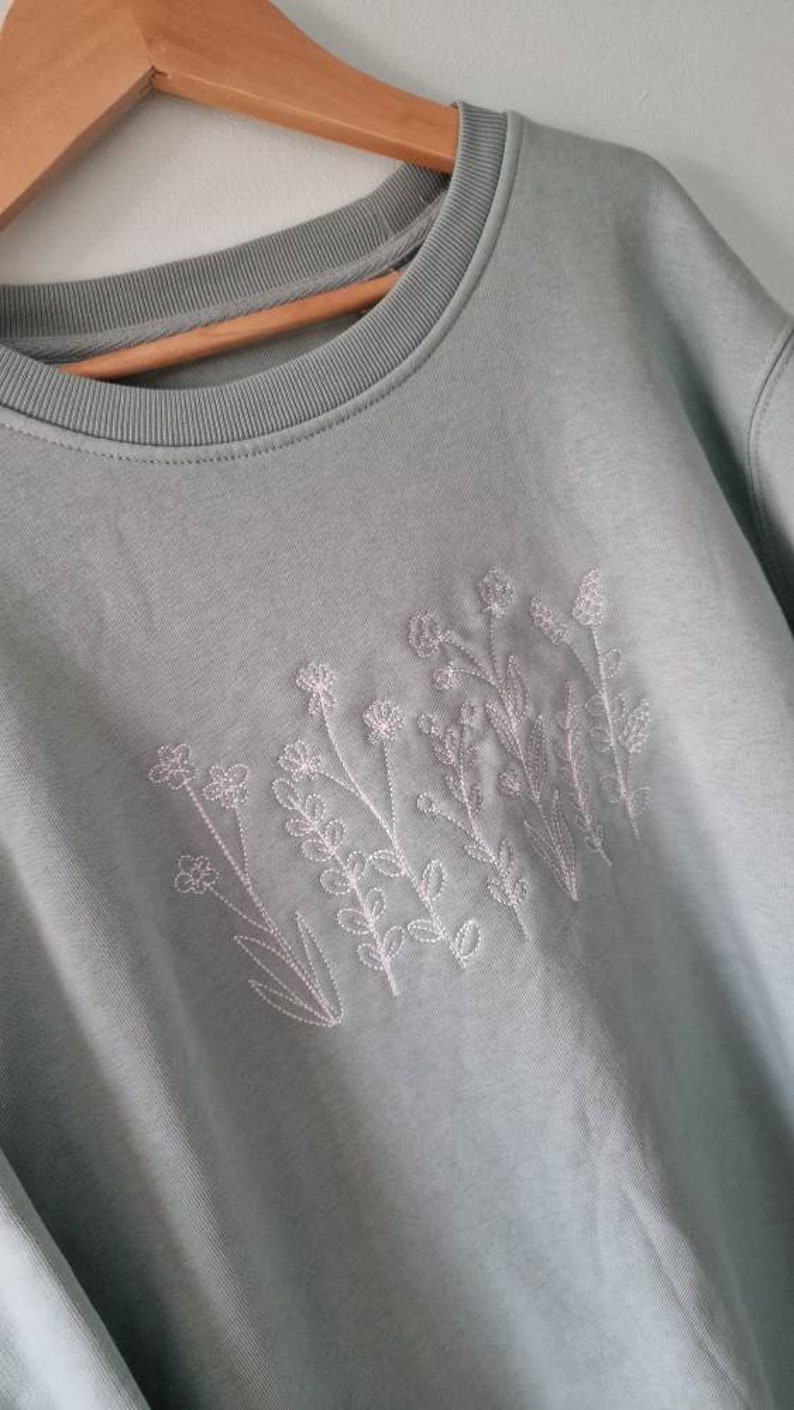 Op de afbeelding: Een lichtblauw sweatshirt met witte geborduurde bloemen.