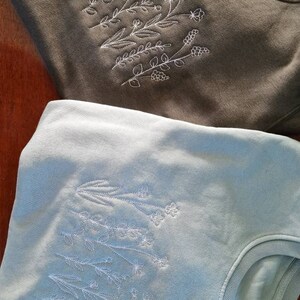 Op de afbeelding: Twee sweatshirts met geborduurde bloemenmotieven. Het bovenste sweatshirt is olijfgroen met witte borduursels. Het onderste sweatshirt is lichtblauw met witte borduursels.