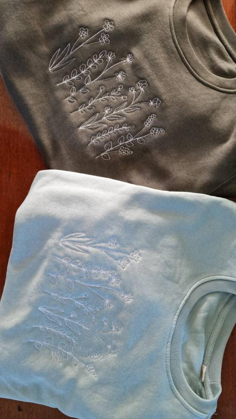 Op de afbeelding: Twee sweatshirts met geborduurde bloemenmotieven. Het bovenste sweatshirt is olijfgroen met witte borduursels. Het onderste sweatshirt is lichtblauw met witte borduursels.