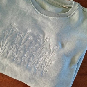 Op de afbeelding: Een lichtblauw sweatshirt met een wit geborduurd bloemenmotief.