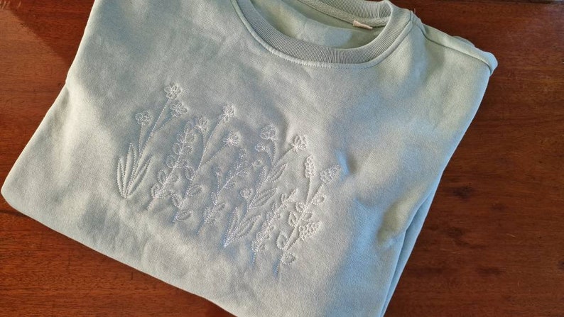 Op de afbeelding: Een lichtblauw sweatshirt met een wit geborduurd bloemenmotief.