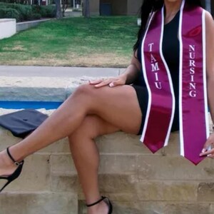 May include: A woman wearing a black dress and a maroon and white graduation sash with the text "TAMU NURSING". She is sitting on a stone wall with a blue water feature behind her.