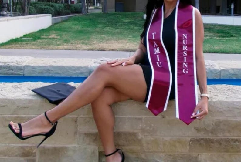 May include: A woman wearing a black dress and a maroon and white graduation sash with the text "TAMU NURSING". She is sitting on a stone wall with a blue water feature behind her.