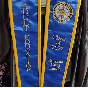 May include: A blue and gold graduation sash with the text "Adult Education" on one side and "Angelo State University Class of 2022 Summa Cum Laude" on the other side.