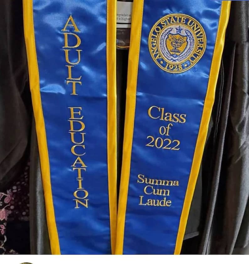 May include: A blue and gold graduation sash with the text "Adult Education" on one side and "Angelo State University Class of 2022 Summa Cum Laude" on the other side.