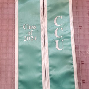 Może przedstawiać: Turkusowa szarfa absolwentka z białymi lam&oacute;wkami i napisem "Class of 2024" po lewej stronie i "CCU" po prawej stronie.