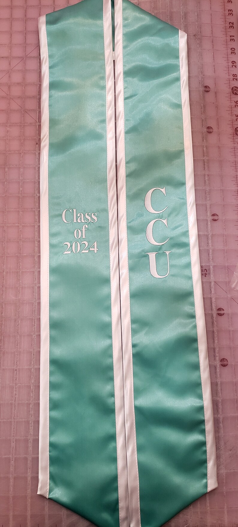 Może przedstawiać: Turkusowa szarfa absolwentka z białymi lam&oacute;wkami i napisem "Class of 2024" po lewej stronie i "CCU" po prawej stronie.